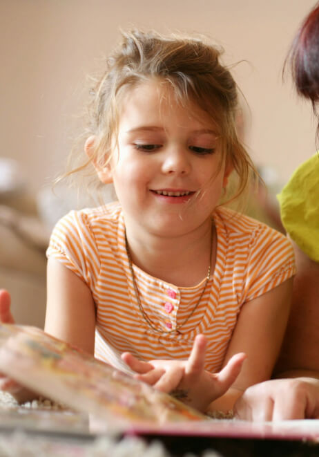 A young girl with blonde hair, wearing an orange striped shirt, smiles while looking at a book. Additionally, a partially visible person in a green shirt is next to her.