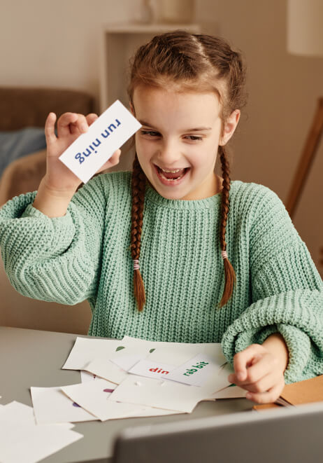 A young girl with braided hair and a green sweater excitedly holds up a flashcard that reads "running" while sitting at a table with more flashcards.