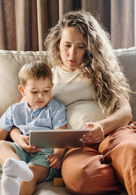 A woman with long hair is sitting on a couch with a young child on her lap. They are both looking at a tablet she is holding.
