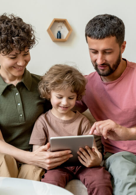Three people sitting on a couch, a child holding a tablet with two adults beside, all looking at the screen and smiling.