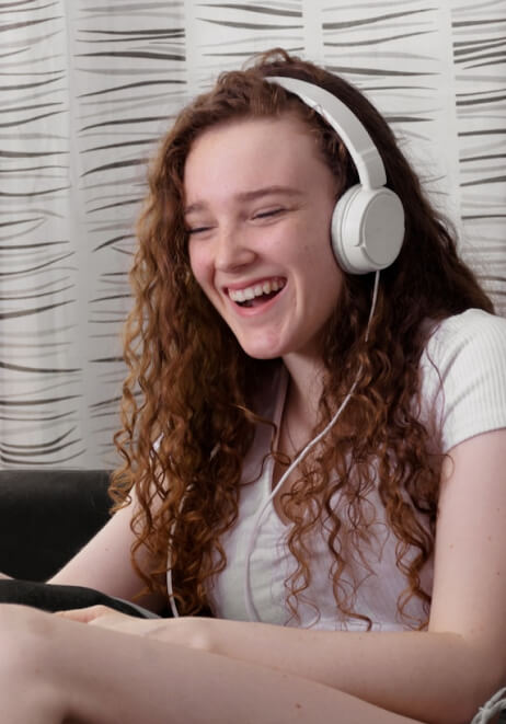A teen wearing white headphones and a white shirt, smiles and laughs while sitting indoors.