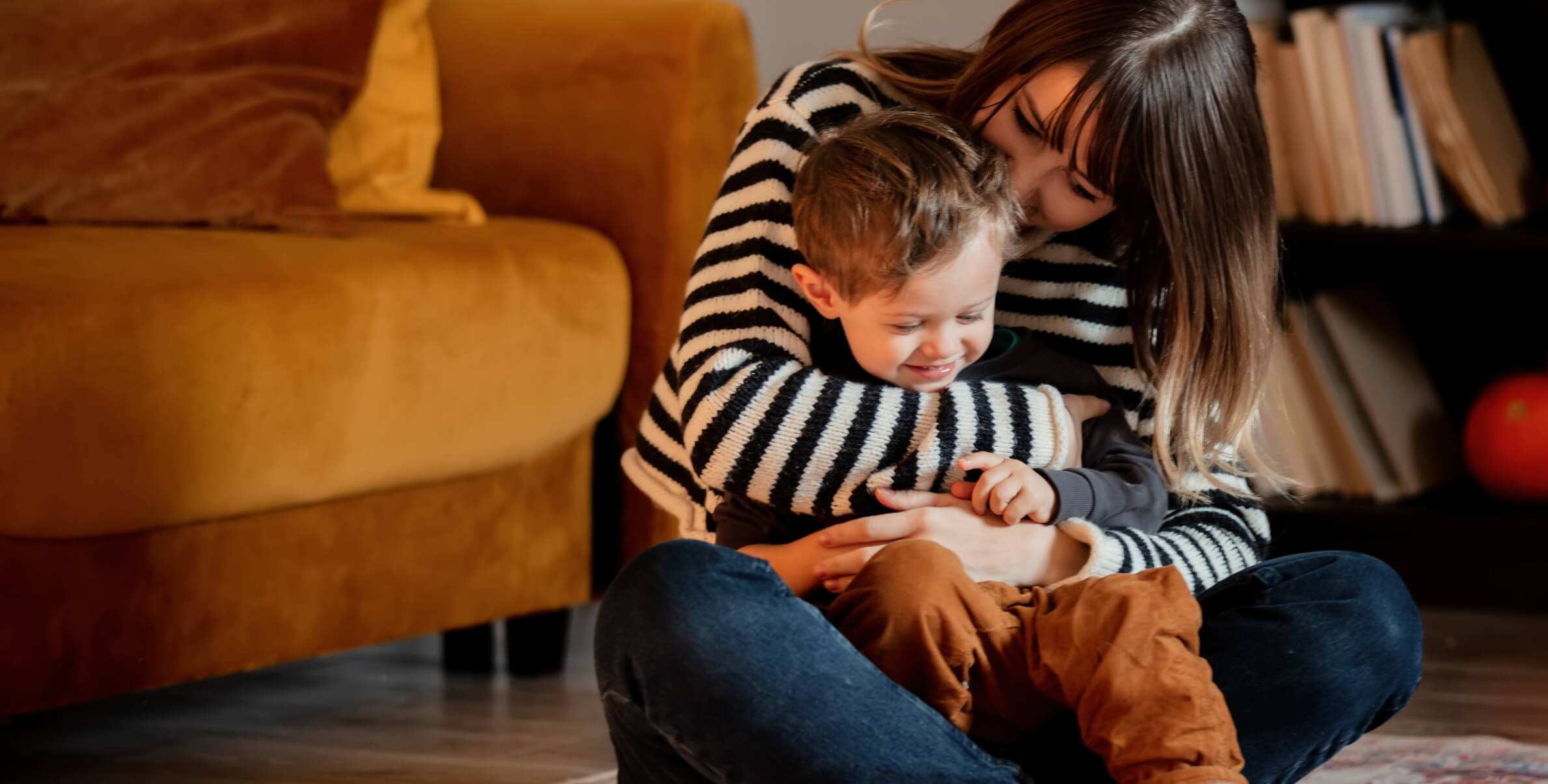 An adult and a child sit together on the floor, with the adult embracing the child. Both appear to be in a cozy living room with a yellow sofa and bookshelf in the background.
