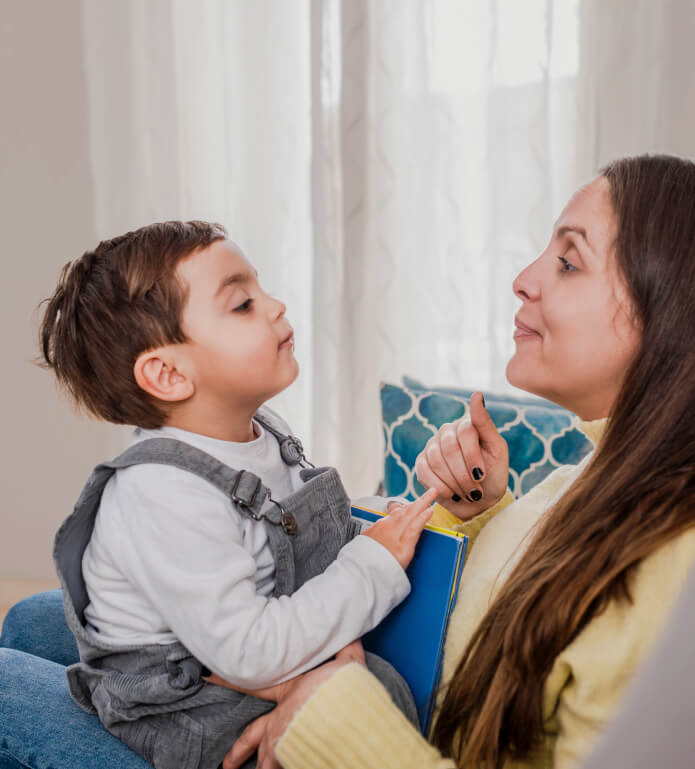 A young boy in overalls sits on a woman's lap, both engaged in conversation. The woman holds a book, and there are patterned cushions and a window with curtains in the background.