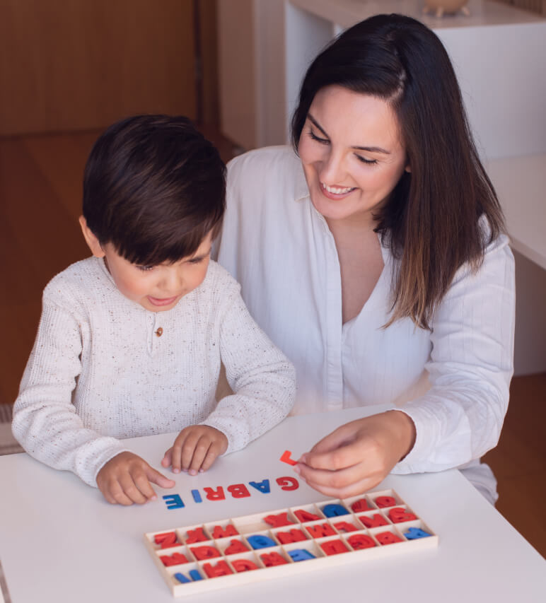An adult and child are sitting at a table, playing with a wooden letter board, spelling out the word "GABRIEL." The adult is smiling and the child seems focused on the activity.