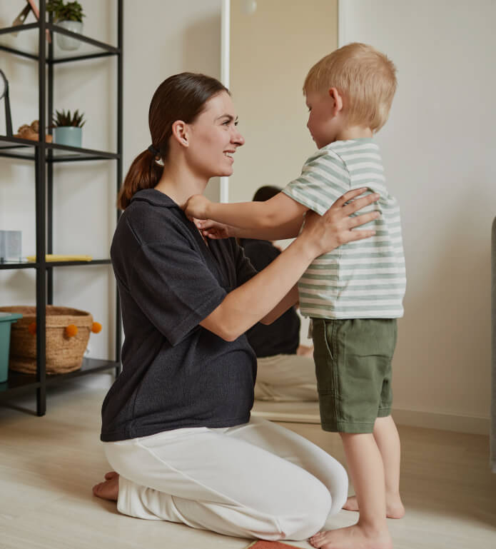 A woman kneels on the floor holding a young boy who is standing in front of her. They are indoors, with shelves and plants in the background.