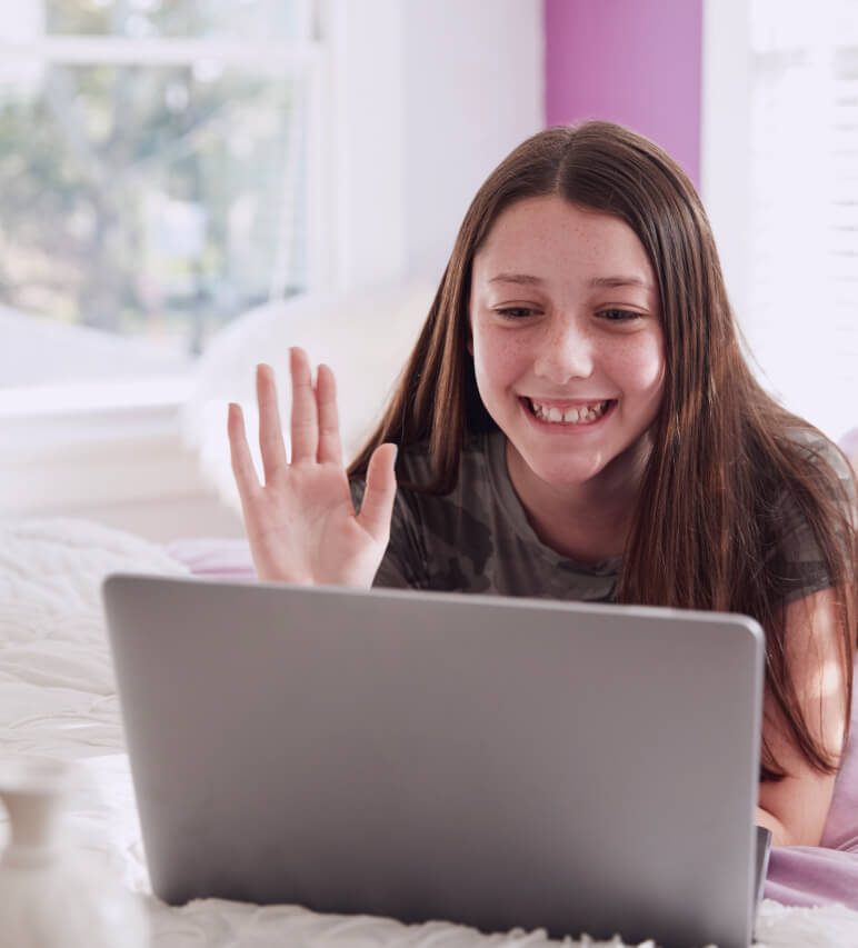 A girl with long brown hair smiles and waves at a laptop screen while lying on a bed in a bright room.