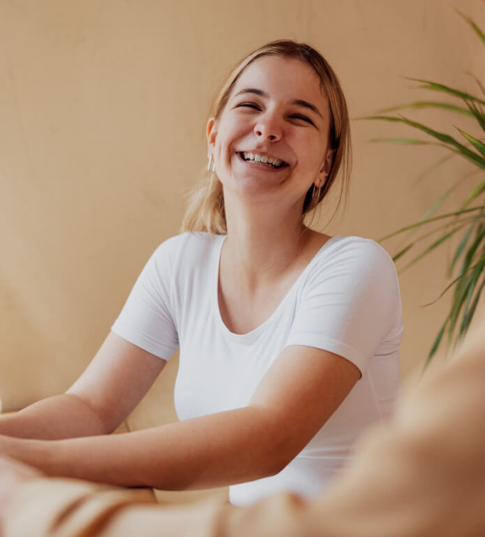 A smiling person wearing a white shirt with blonde hair tied back sits indoors next to a plant.