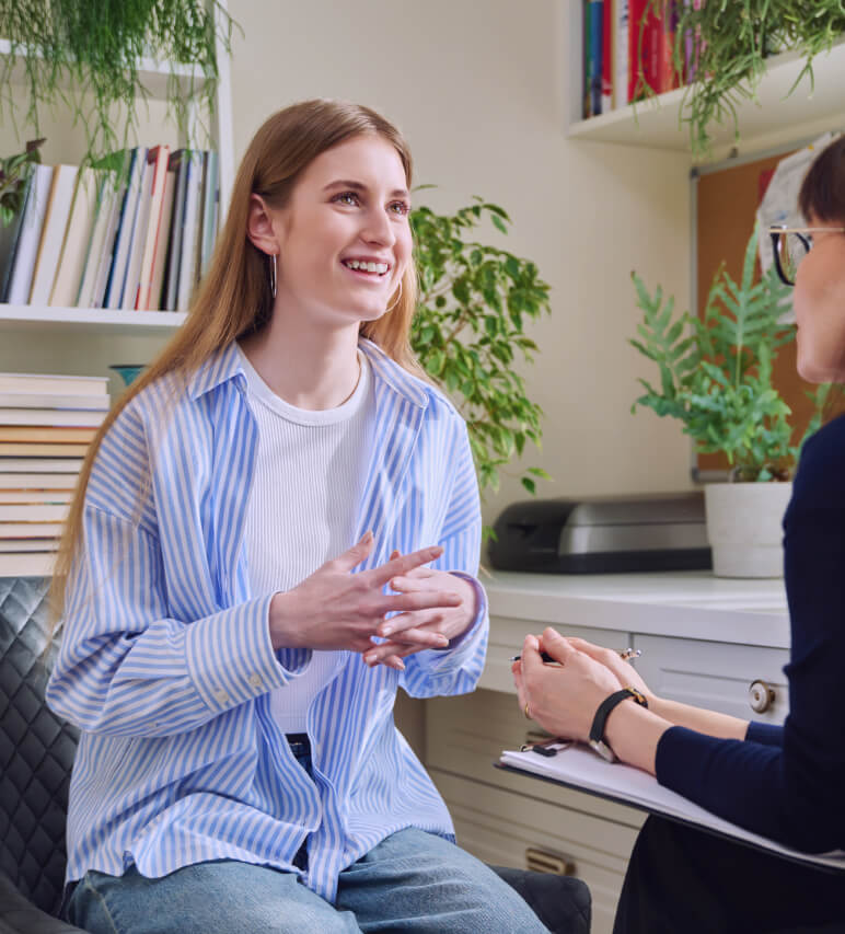 A young woman sits talking with a seated professional holding a clipboard in an office with shelves, books, and plants.
