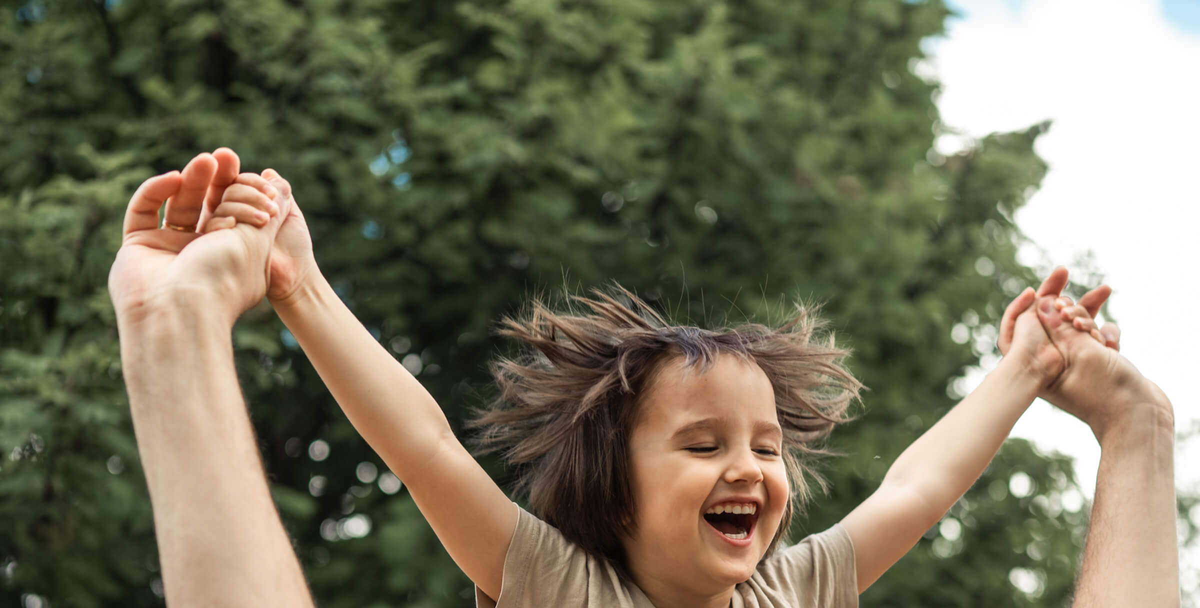 A child being lifted in the air by adult hands, smiling with trees in the background.