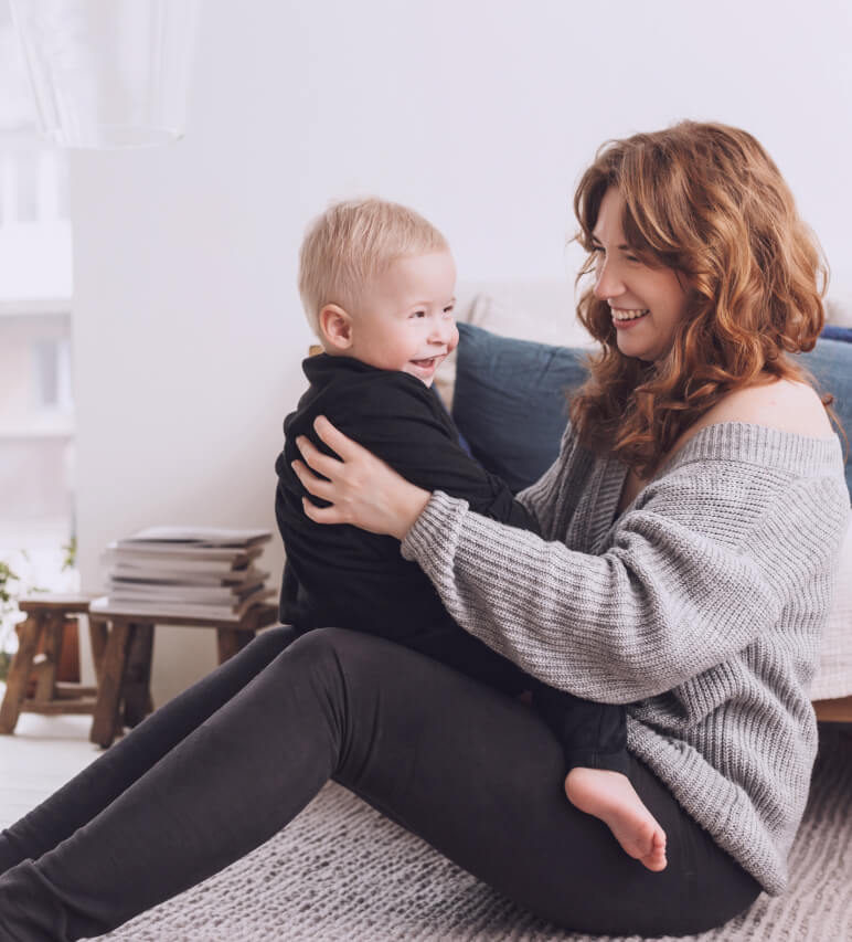 A woman with curly hair, wearing a grey sweater, sits on the floor holding a smiling child dressed in black. They are in a cozy, well-lit room with a stack of books in the background.
