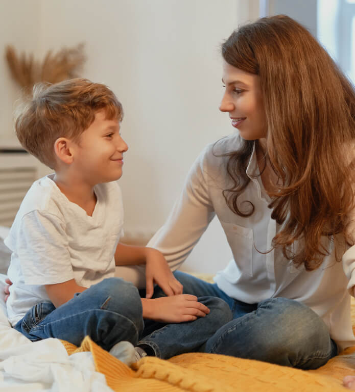 A woman and a young boy, both with brown hair and wearing casual clothes, sit on a bed, smiling and looking at each other.