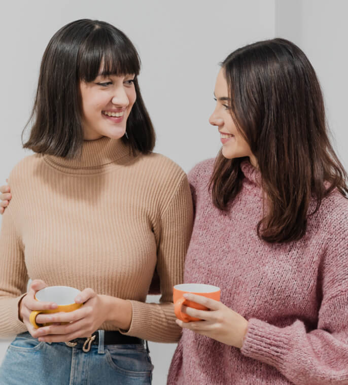 Two women, one in a beige turtleneck and the other in a pink sweater, smile at each other while holding ceramic mugs.