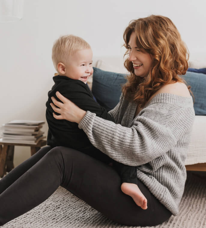 A woman with wavy hair wearing a gray sweater sits on the floor, holding a smiling toddler dressed in black. They are sitting in a cozy room with a stack of books in the background.
