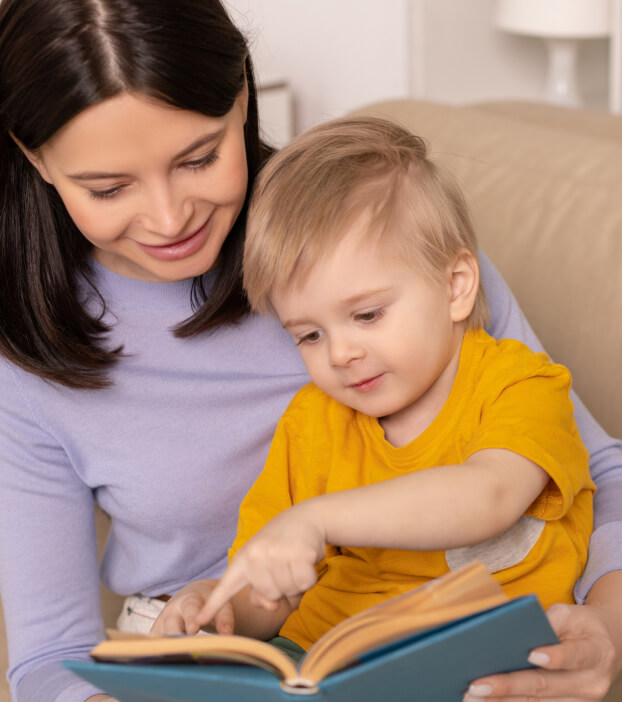 An adult and a child are sitting on a couch reading a book together. The child is pointing at the book while the adult smiles and watches.