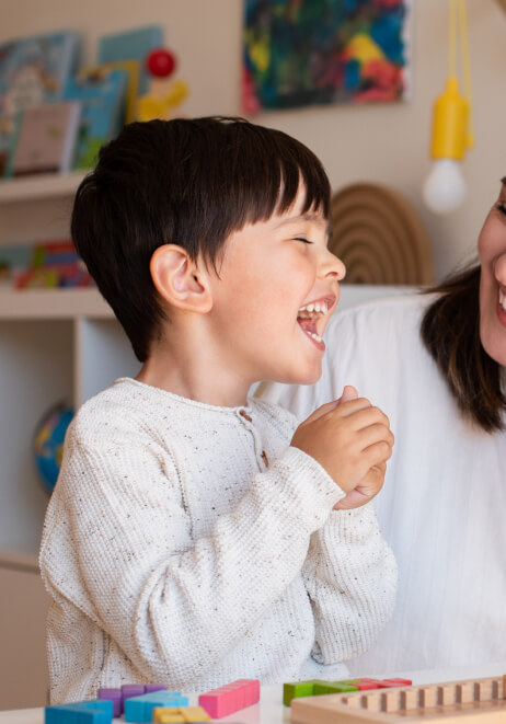A young child with dark hair laughs joyfully, wearing a cream-colored sweater. An adult partially visible beside them shares in the moment. Shelves with books and toys are in the background.