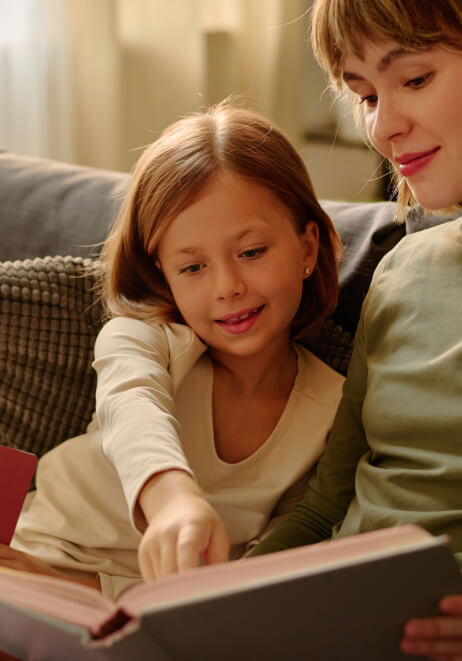Speech therapist and a child reading a book together on a couch. The child is pointing at a page, and they both appear engaged in the activity.