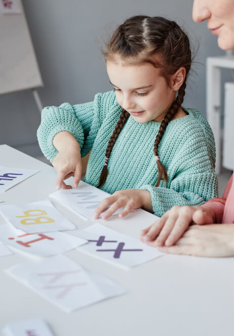 A young girl with braided hair cuts paper with letters on it, while an speech pathologist watches beside her at a white table.