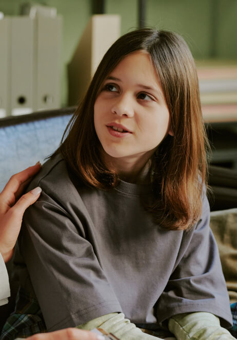 A young, brown-haired girl is sitting indoors on a couch, looking slightly to the side and talking. A speech therapist's hand is gently placed on her shoulder.