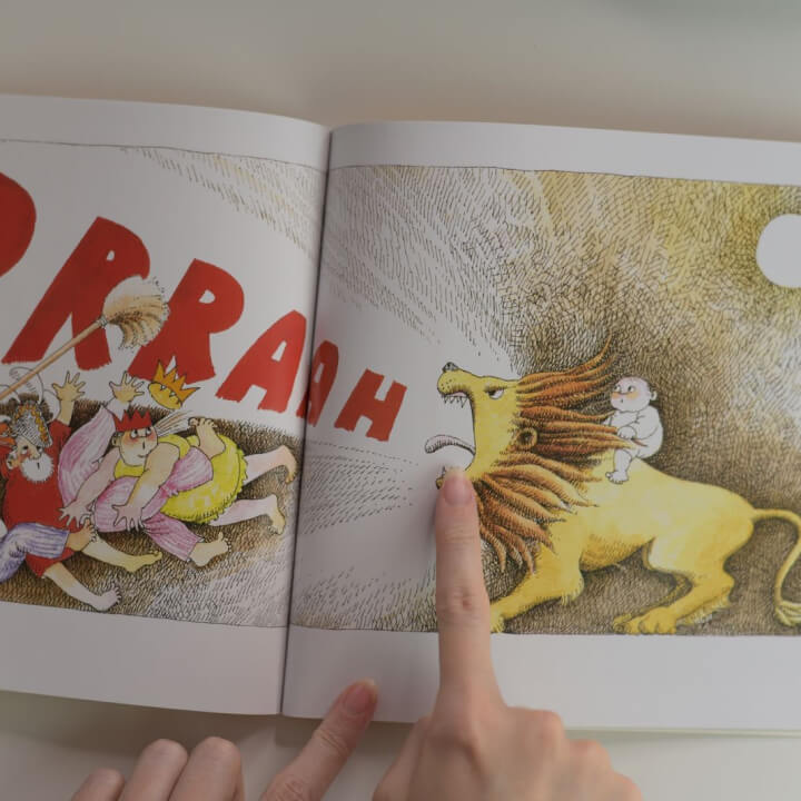 A person points to an illustration in a book showing a lion with a child riding it. Two characters run away, and red letters spell "PRRAH" in the background.