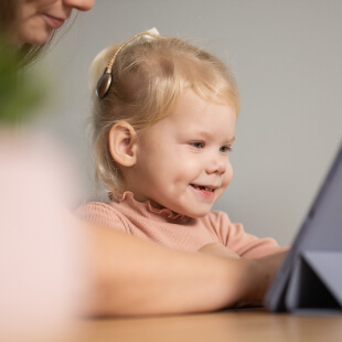 A young child, wearing a hearing aid, smiles while looking at a tablet with an adult nearby.