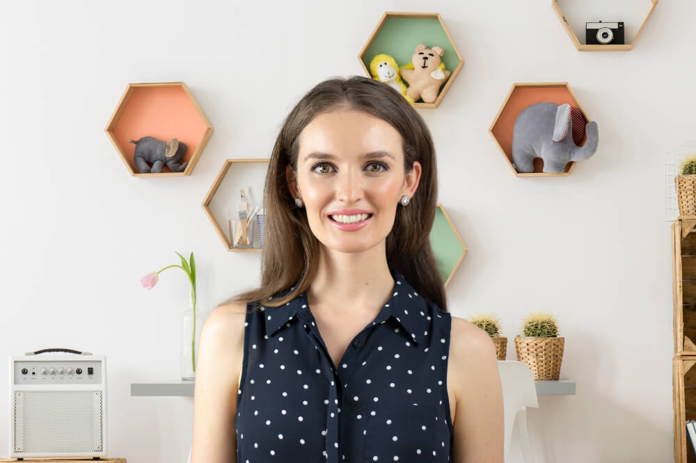 Lauren Crumlish is standing in a room with hexagonal shelves on the wall behind her, displaying various items like stuffed animals and a camera. She is wearing a navy blue polka dot sleeveless top.