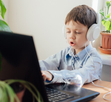 A young boy wearing headphones uses a laptop at a desk. He is focused on the screen, and there are potted plants in the background.