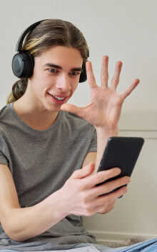 A person with long hair wearing headphones smiles and waves at a smartphone screen while holding the device in their other hand.