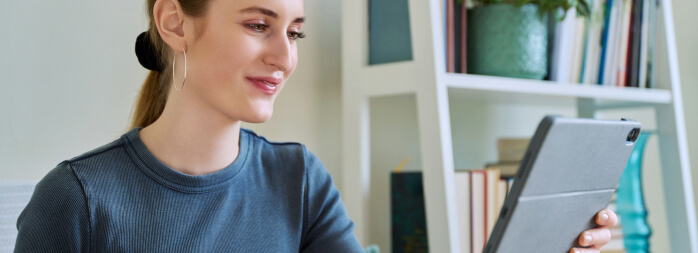 A woman is smiling while looking at a tablet indoors, with a bookshelf in the background.