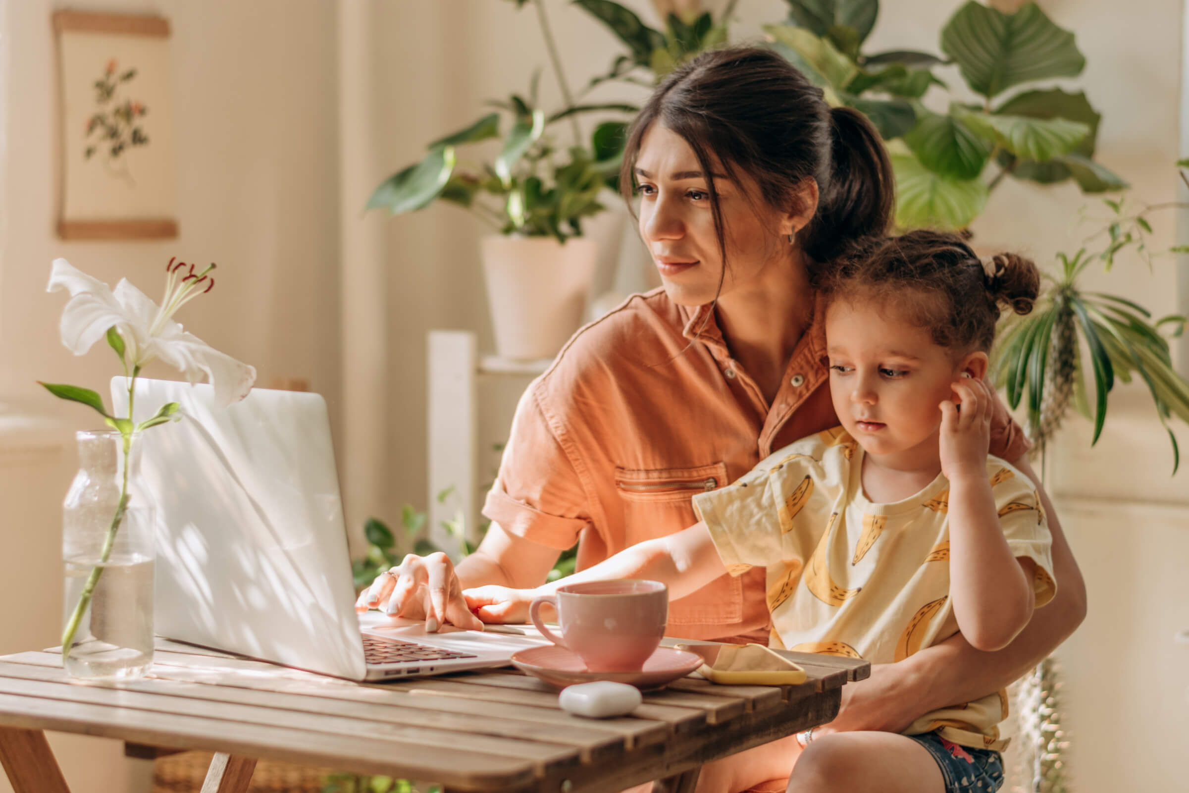 Mother works on a laptop with a child sitting on her lap at a wooden table, surrounded by potted plants and a vase of flowers.