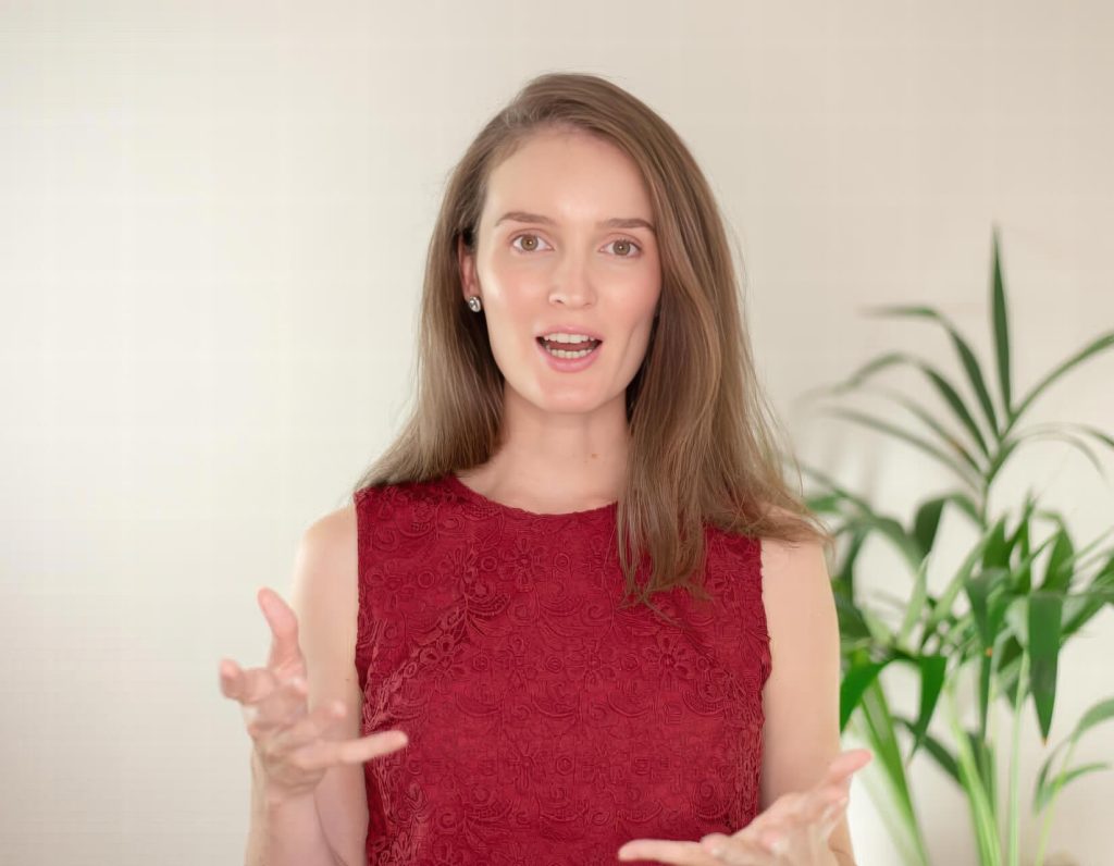 A woman in a red lace sleeveless top is speaking with expressive hand gestures. A green plant is in the blurred background.