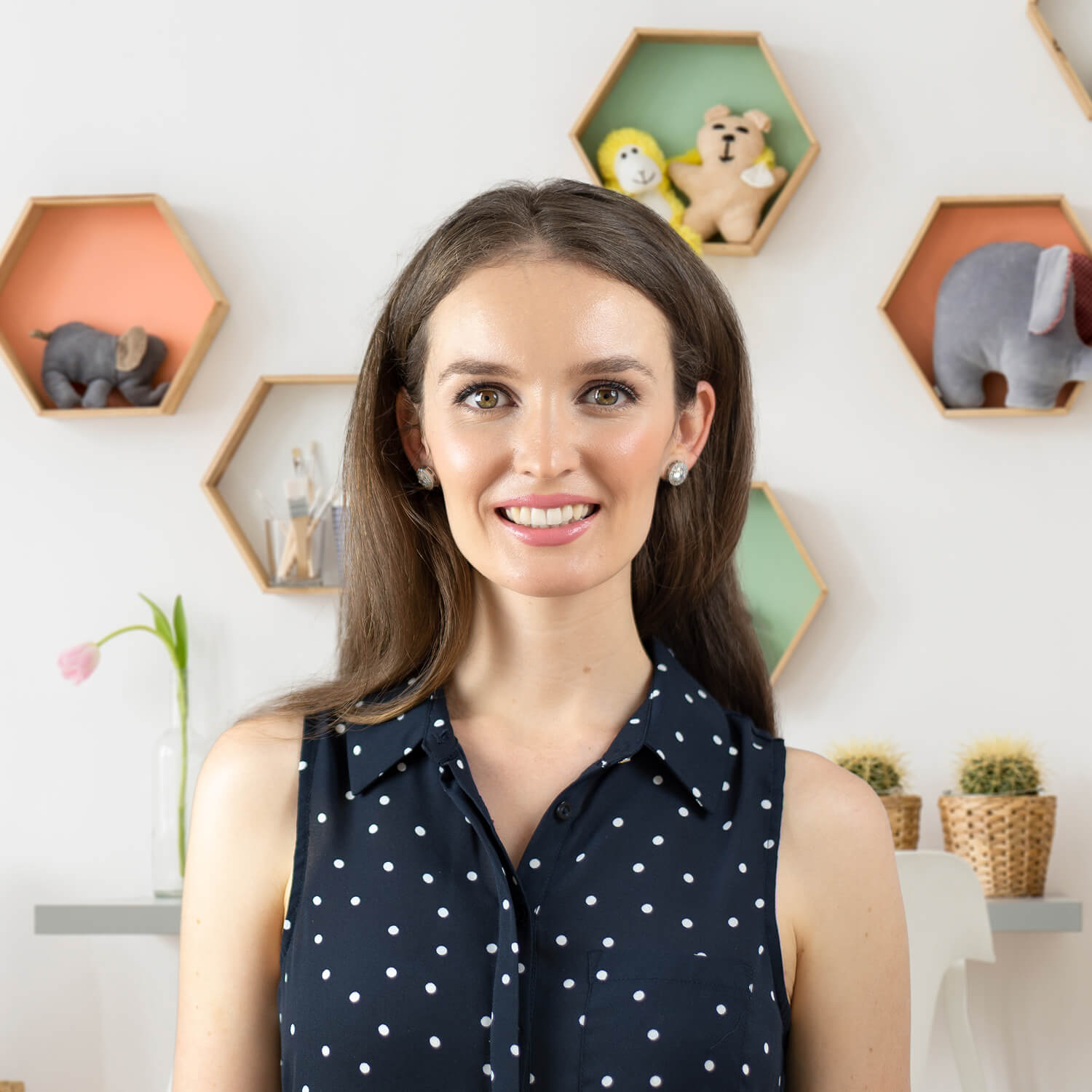 Dr Lauren Crumlish smiling in front of hexagonal shelves with toys and decor; wearing a sleeveless polka dot blouse.