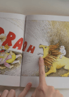 A person points to an illustration in a children's book depicting a roaring lion with a rider on its back and people running away.