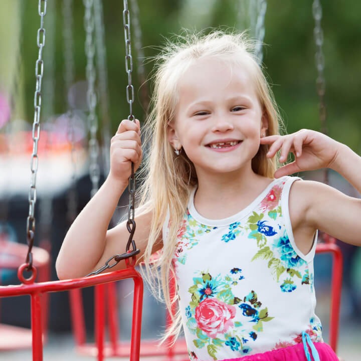 A young girl with long blonde hair smiles while standing next to a red swing, wearing a white floral dress.